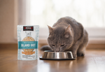 A gray cat eating from a metal bowl next to a pouch of KOHA Bland Diet canned cat food on a wooden floor.