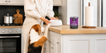 A person in a cream striped shirt and pants stands in a bright kitchen with a beagle standing on its hind legs at the counter, reaching for a white bowl. A can of KOHA Limited Ingredient dog food with a purple label is on the wooden countertop next to a paper towel holder.