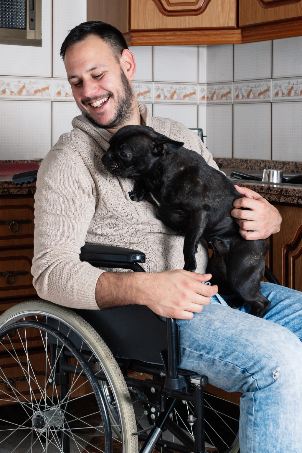 A smiling man in a wheelchair wearing a beige knit sweater and blue jeans holds a small black dog in his lap in a kitchen setting.