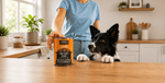 A person in a blue shirt stands in a bright kitchen behind a dog, holding a can of KOHA Big Easy Feast dog food on a wooden countertop.