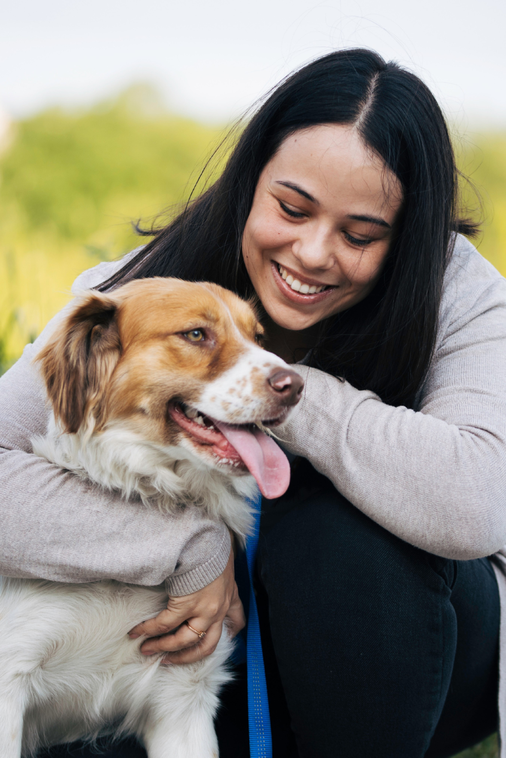 A woman smiling and hugging a brown and white dog outdoors, wearing a light gray long-sleeve top and dark pants.